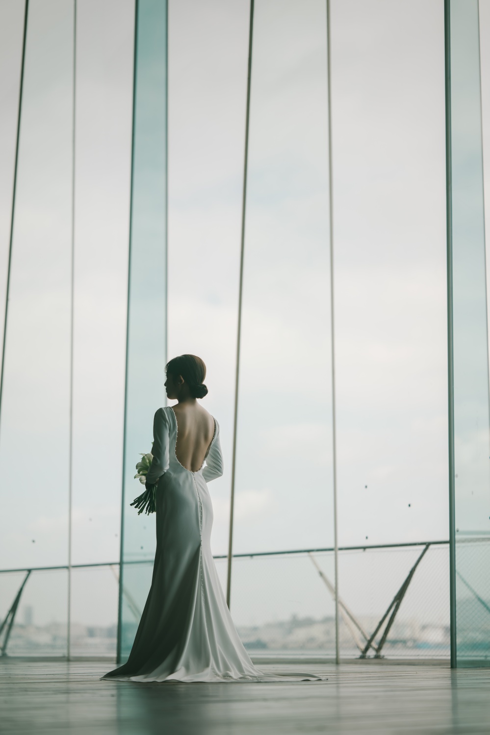 大きなガラス窓から光が差し込むロケーションにて、パールのあしらわれた美しいシンプルなウェディングドレスを纏う花嫁様のバックスタイル