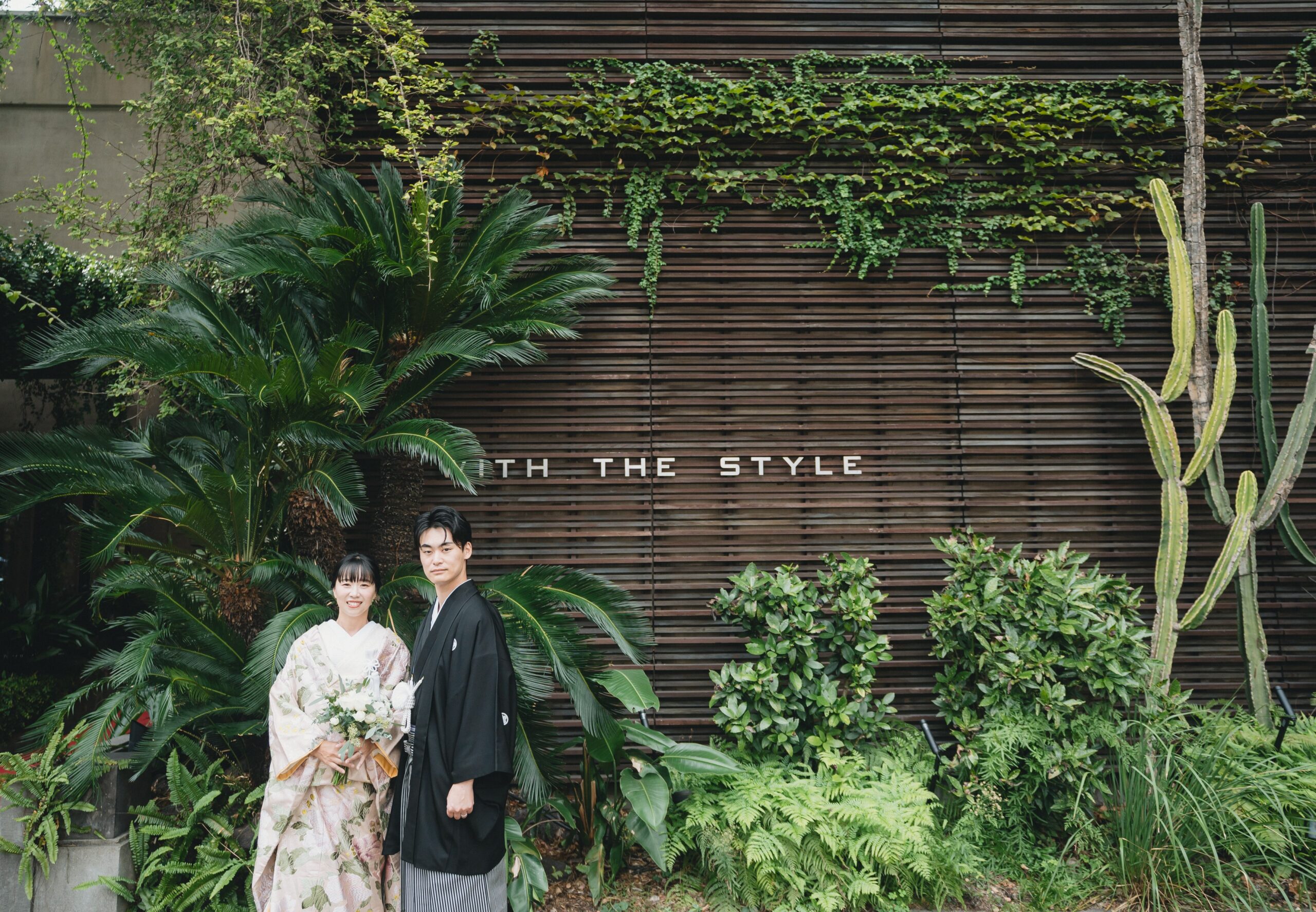 大人でセンスフルなウィズ ザ スタイル フクオカの正面玄関でのご新郎様ご新婦様のお写真。ご新婦様は桃色の色打掛と、グリーンとホワイトのブーケをコーディネート。