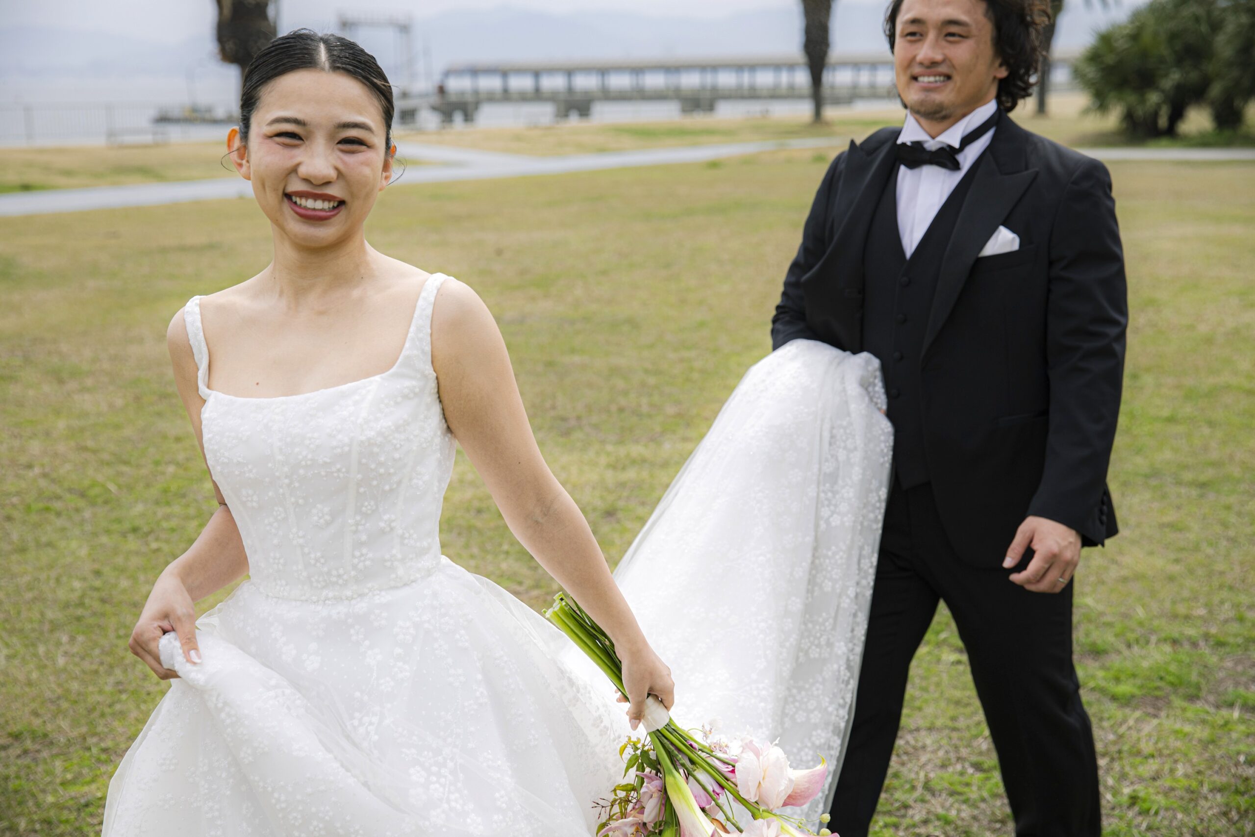 トリート福岡の提携会場、ザ ルイガンズ スパ&リゾートのガーデンをウェディングドレスのトレーンを持って歩くご新郎様とカメラ目線で楽しそうに笑顔を浮かべるご新婦様。