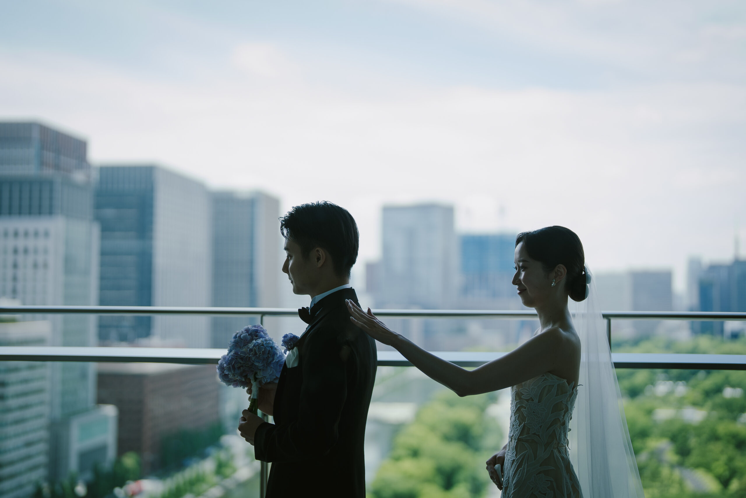 皇居の美しい緑をバックにトリートのオスカーデラレンタのウェディングドレスを纏い、ブラックタキシードに身を包むご新郎様の肩をたたくご新婦様の様子
